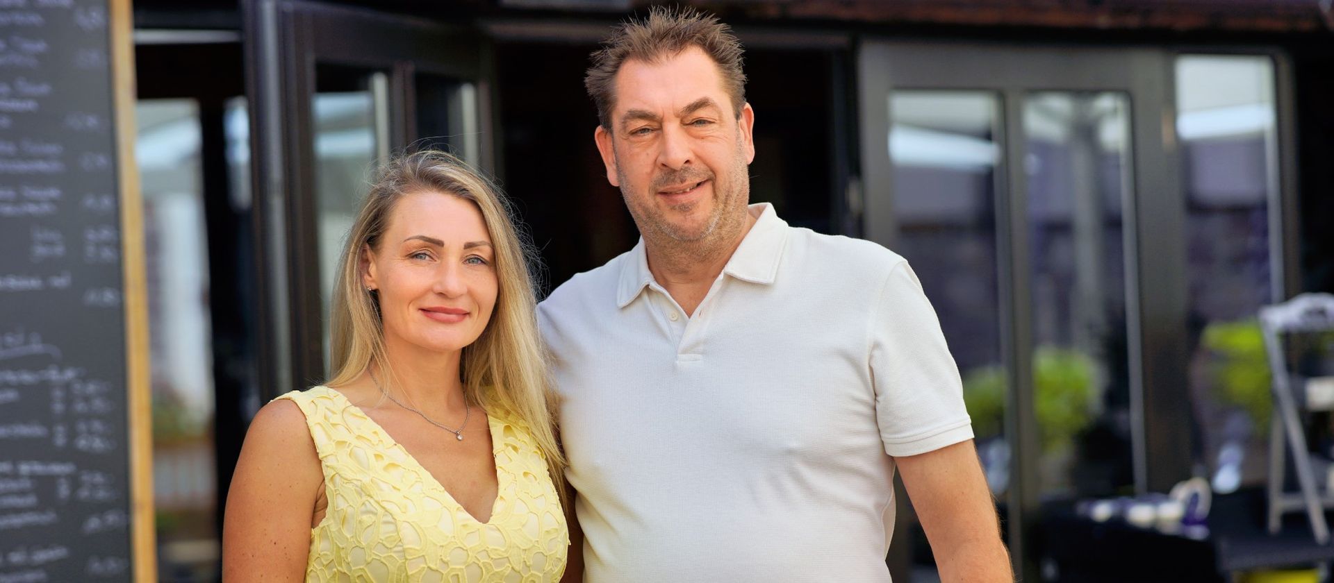 Ein Mann und eine Frau stehen vor einem Restaurant. Die Frau trägt ein gelbes Kleid und der Mann ein weißes Hemd.