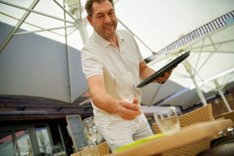 Lächelnder Kellner im weißen Hemd, der unter offenen Sonnenschirmen dem Tisch die Speisekarte reicht.