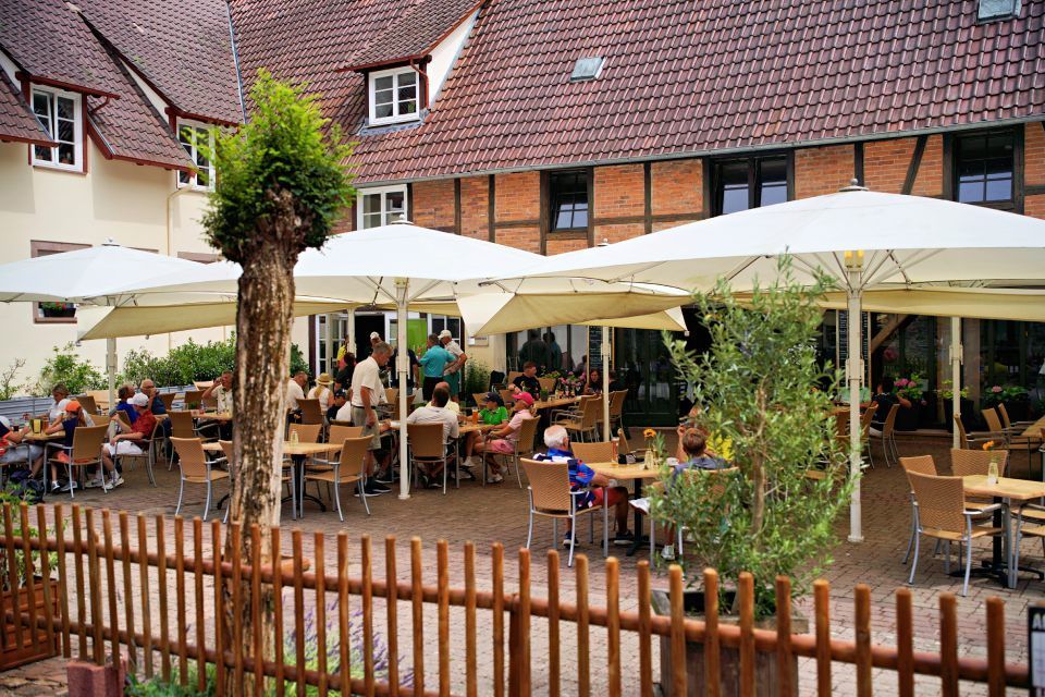 Restaurant im Freien mit Gästen unter Sonnenschirmen; Tische, Stühle und Gebäude im Hintergrund.