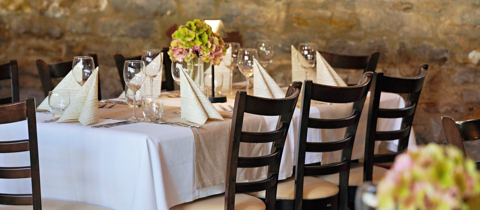 Ein für eine formelle Veranstaltung gedeckter Esstisch mit weißer Tischwäsche, Servietten und Blumenschmuck in einem Raum mit Steinwänden.