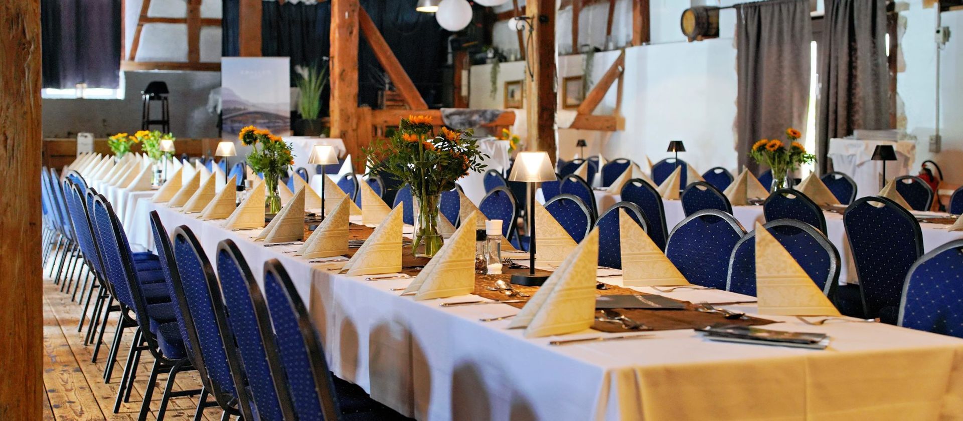 Lange, für ein Bankett gedeckte Esstische mit blauen Stühlen, weißen Tischdecken und Blumengestecken.