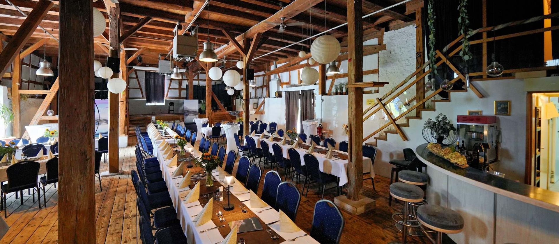Großer Saal mit langen, für ein Bankett gedeckten Tischen, rustikalen Holzbalken, Bar und Treppe.