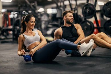 Ein Strauß rosa und roter Blumen mit einem Etikett mit der Aufschrift „Schöne Pfingsten“. Frau und Mann machen im Fitnessstudio Rumpftraining mit Kettlebells.