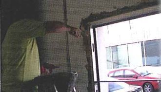 Un hombre está trabajando en una puerta de garaje con un coche rojo aparcado al fondo.