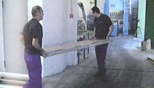 Dos hombres transportan un gran trozo de madera en un almacén.