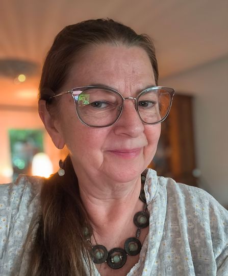 Frau mit Brille und Perlenkette lächelt sanft im warmen Licht eines Innenraums.