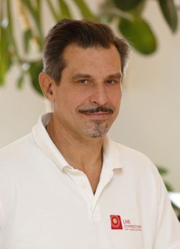 Ein Mann in einem weißen Poloshirt steht in einem Innenraum; im Hintergrund sind verschwommene grüne Blätter zu sehen.