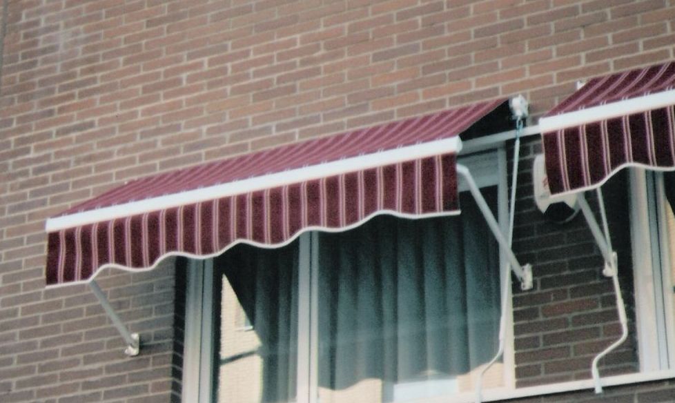 Toldo de rayas rojas y blancas sobre una ventana de un edificio de ladrillo, sostenido por soportes de metal blanco.