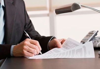 Persona vestida de traje firmando un documento en un escritorio con un bolígrafo.