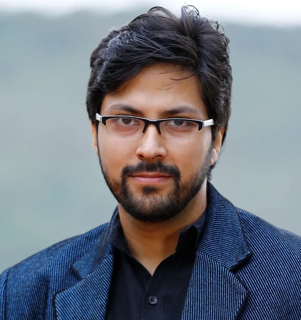 Head-and-shoulders portrait of a man with glasses and a short beard wearing a blue jacket outdoors