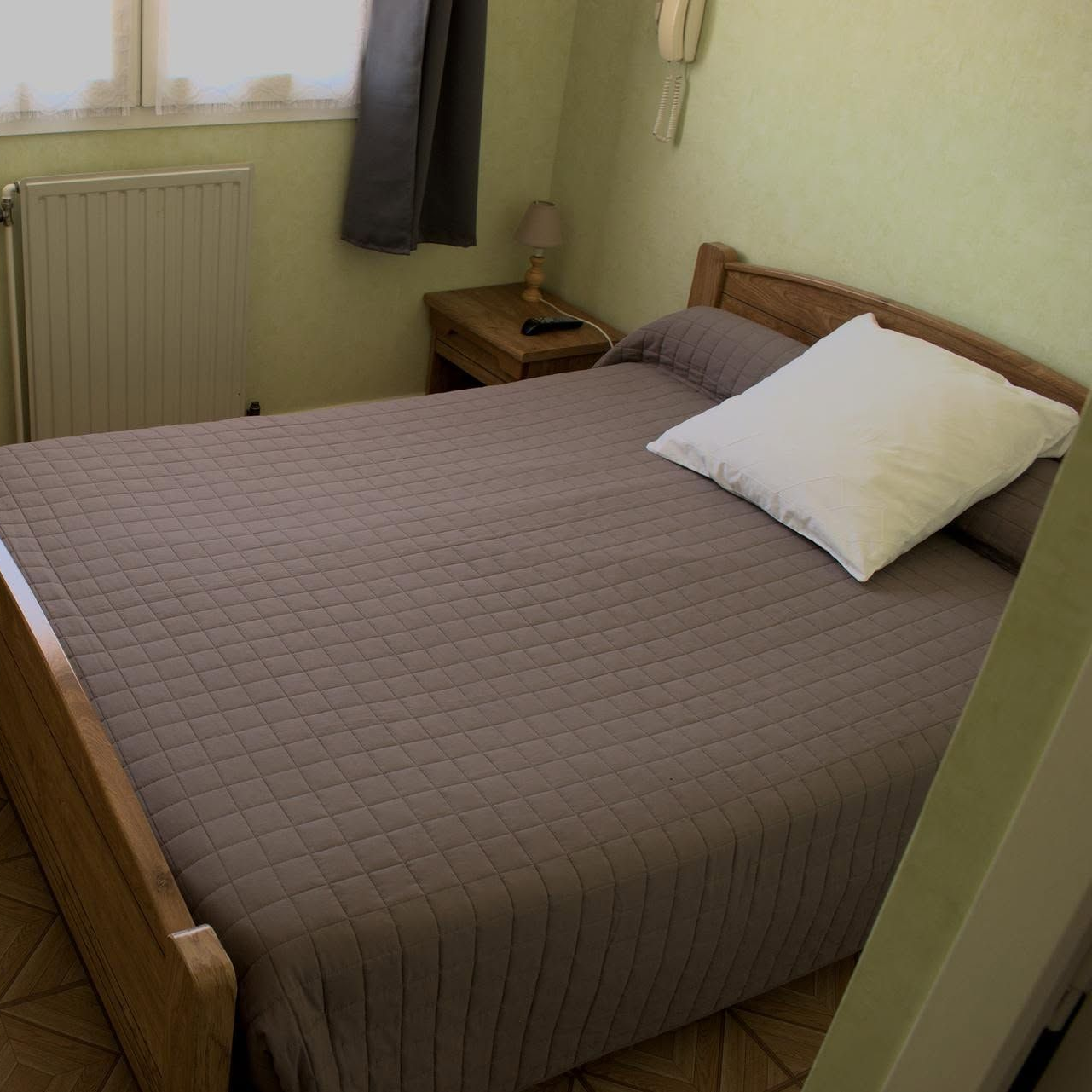 Petite chambre avec un lit fait, une dessus de lit marron, un oreiller blanc et une table de chevet près d'une fenêtre.