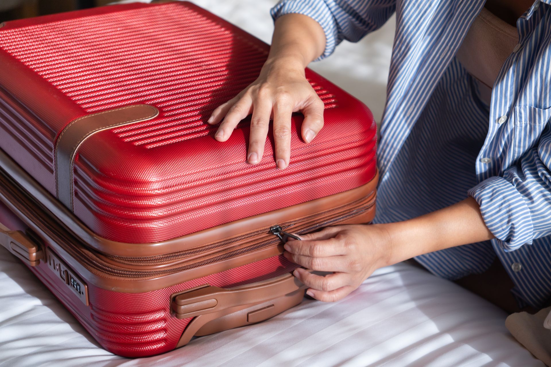 Des mains qui ferment une fermeture éclair marron sur une valise rigide rouge.
