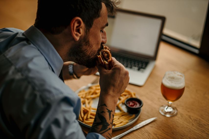 Une personne mange un sandwich à une table avec des frites, une boisson à côté d'un ordinateur portable ouvert.