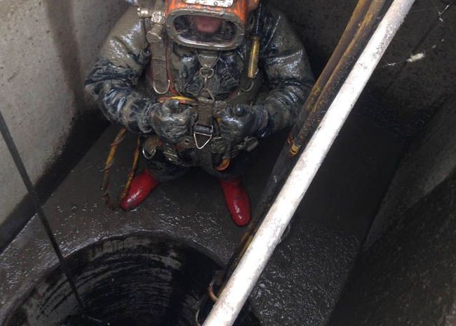 Plongeur en casque et combinaison assis dans un puits étroit et humide, chaussé de bottes rouges et muni d'un tuyau.