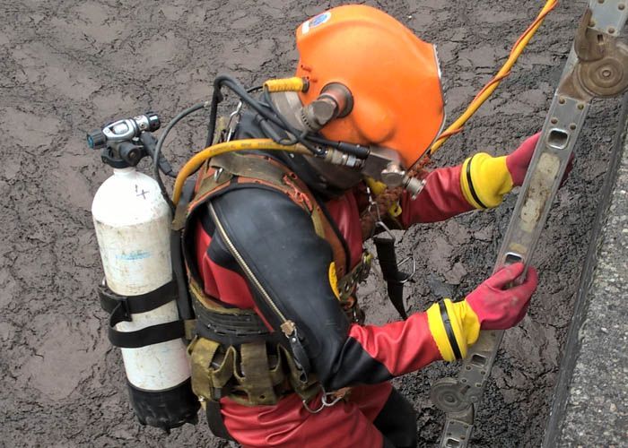 Un ouvrier portant un casque orange et un équipement de sécurité escalade une paroi rocheuse à l'aide de cordes et d'une bonbonne d'eau sur le dos.