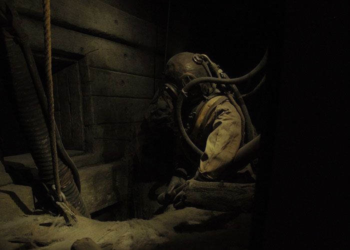 Un scaphandrier dans un tunnel faiblement éclairé, accroupi contre un mur dans un environnement industriel sombre.
