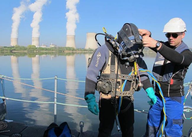 Deux plongeurs sous-marins préparent leur matériel sur un bateau au bord d'une rivière, avec des tours de refroidissement en arrière-plan.