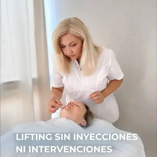 Una mujer con una bata blanca realiza un procedimiento de lifting sin inyecciones en el rostro de otra mujer en una habitación blanca.