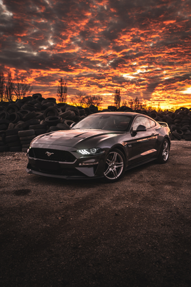 Une mustang noire est garée devant un tas de pneus au coucher du soleil.