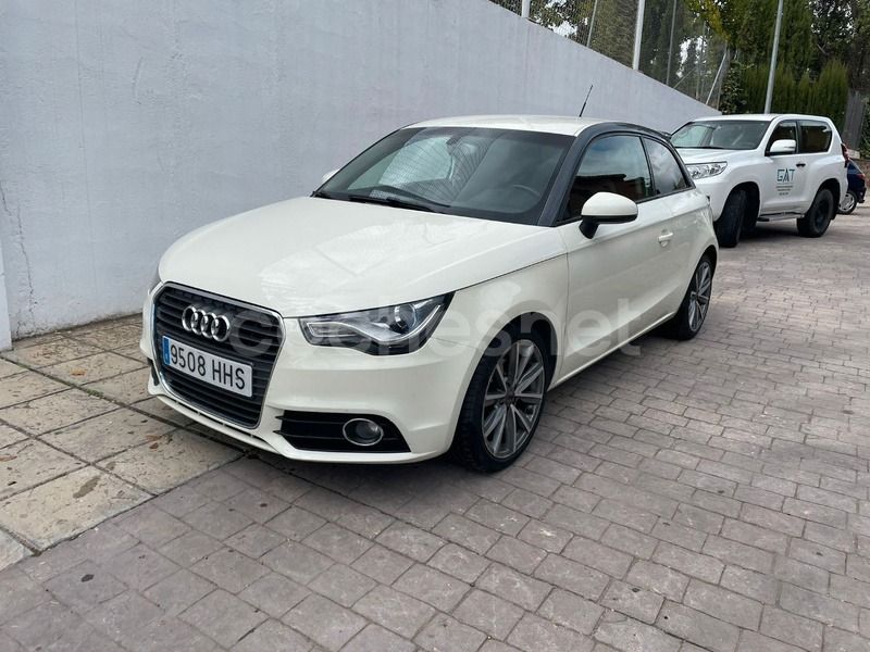Un Audi A1 hatchback blanco de tres puertas estacionado en un aparcamiento pavimentado, con un SUV blanco al fondo.