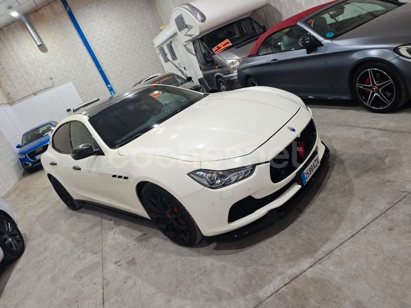 Un Maserati Ghibli blanco estacionado en un almacén, con un descapotable gris y un coche azul parcialmente visibles cerca.