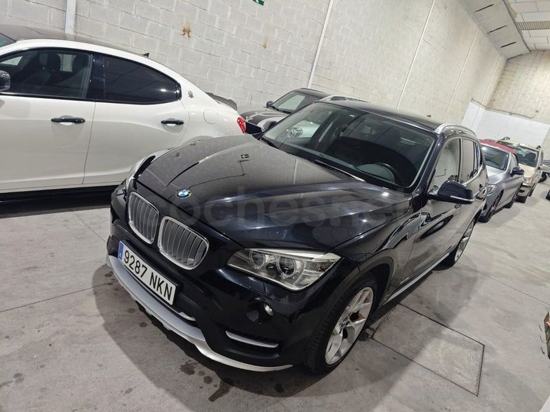 Un SUV BMW X1 negro estacionado en un garaje, colocado en un ligero ángulo cerca de un coche blanco y otros vehículos.