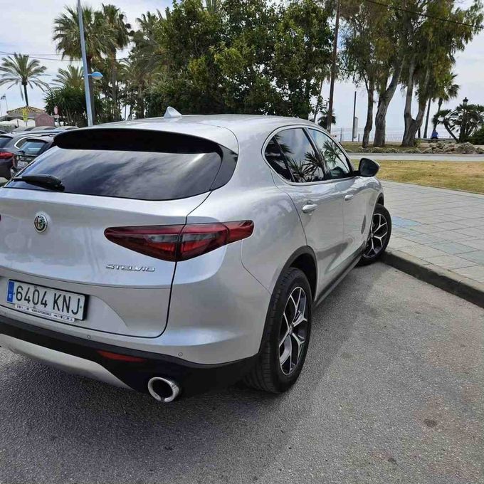 Un SUV Alfa Romeo Stelvio plateado estacionado en un terreno pavimentado con palmeras.
