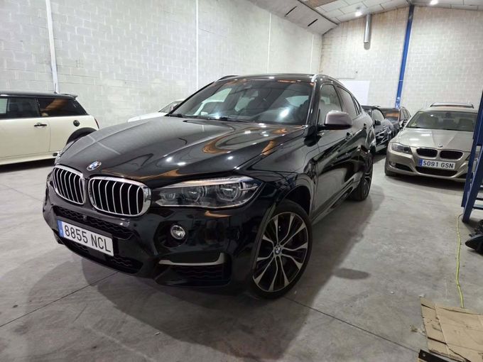 Un SUV BMW X6 negro estacionado dentro de un gran garaje de estilo industrial, con otros vehículos visibles al fondo.