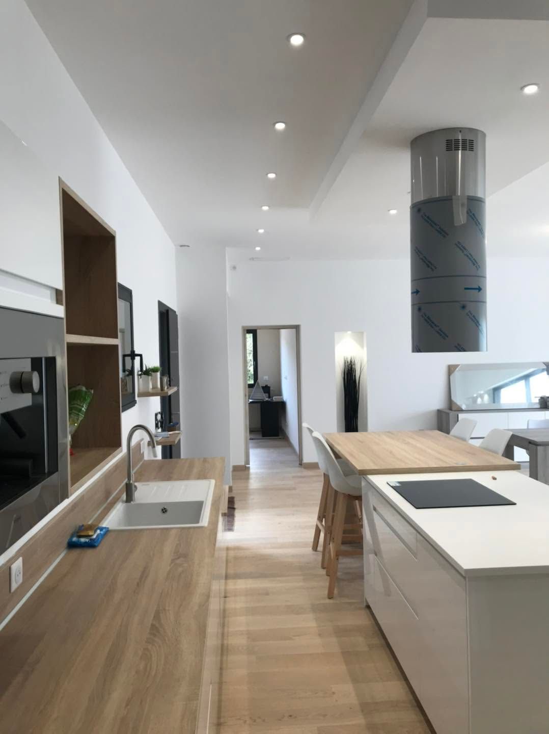 Cuisine blanche moderne avec comptoirs en bois, îlot, évier et éclairage encastré au plafond, vue depuis un couloir.