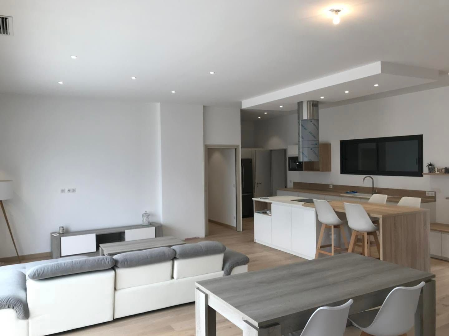 Salon et cuisine modernes et lumineux à aire ouverte, avec meubles blancs et en bois, canapé gris et table à manger.