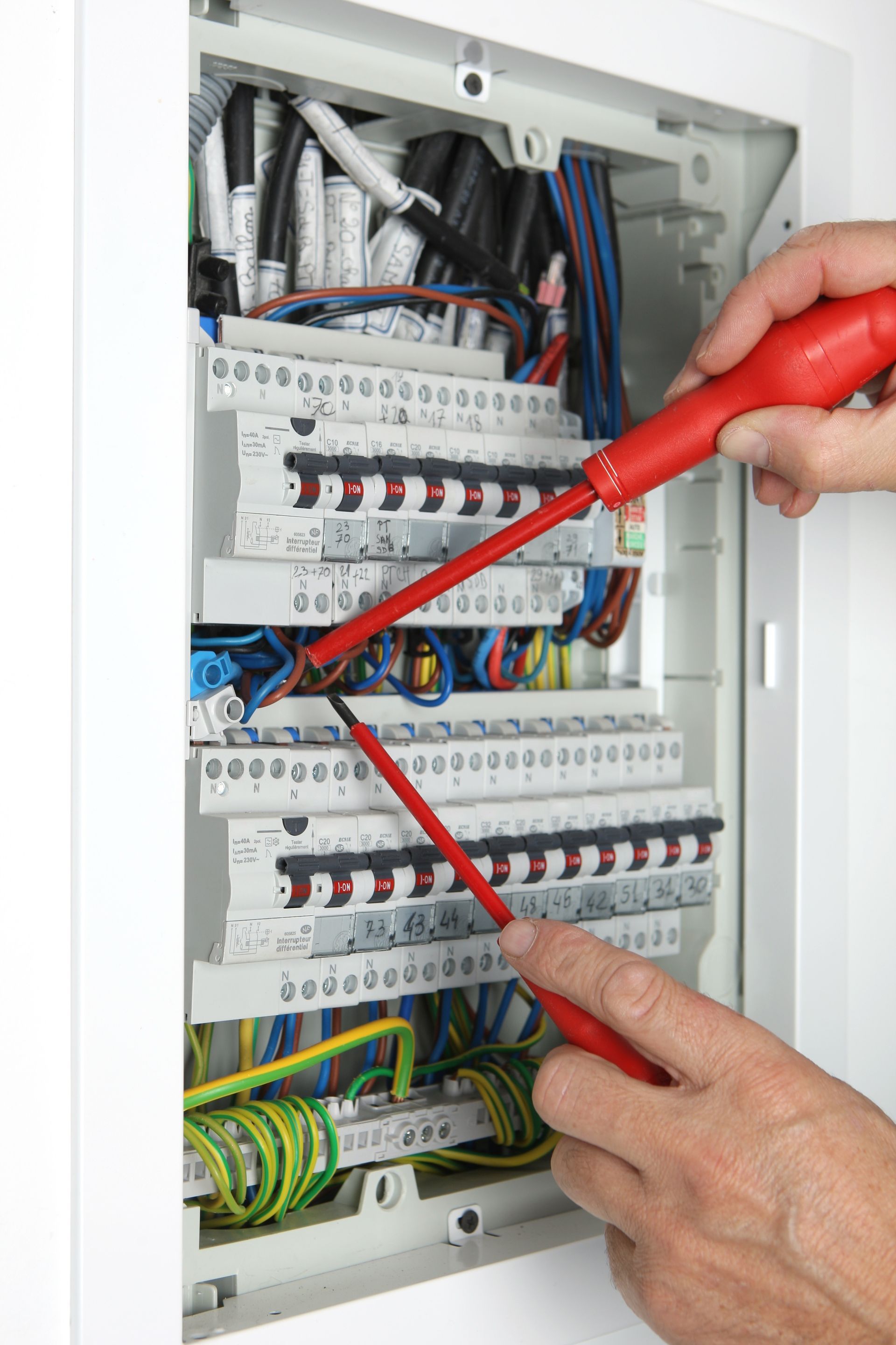 Des mains utilisent des sondes rouges de multimètre sur un panneau de disjoncteurs électrique ouvert, avec des câbles.