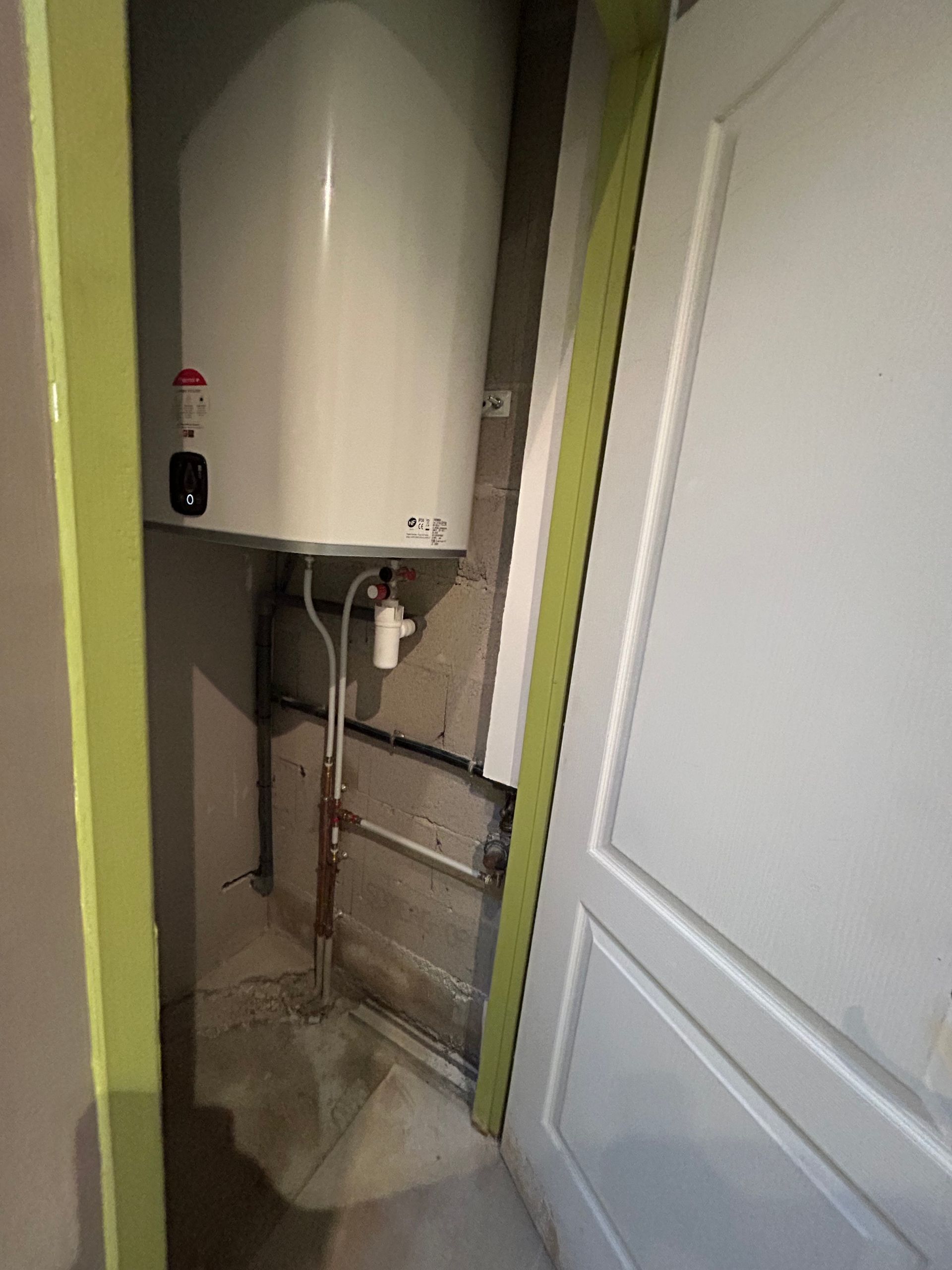 Placard utilitaire étroit avec un chauffe-eau blanc, des tuyaux apparents et une porte blanche entrouverte.