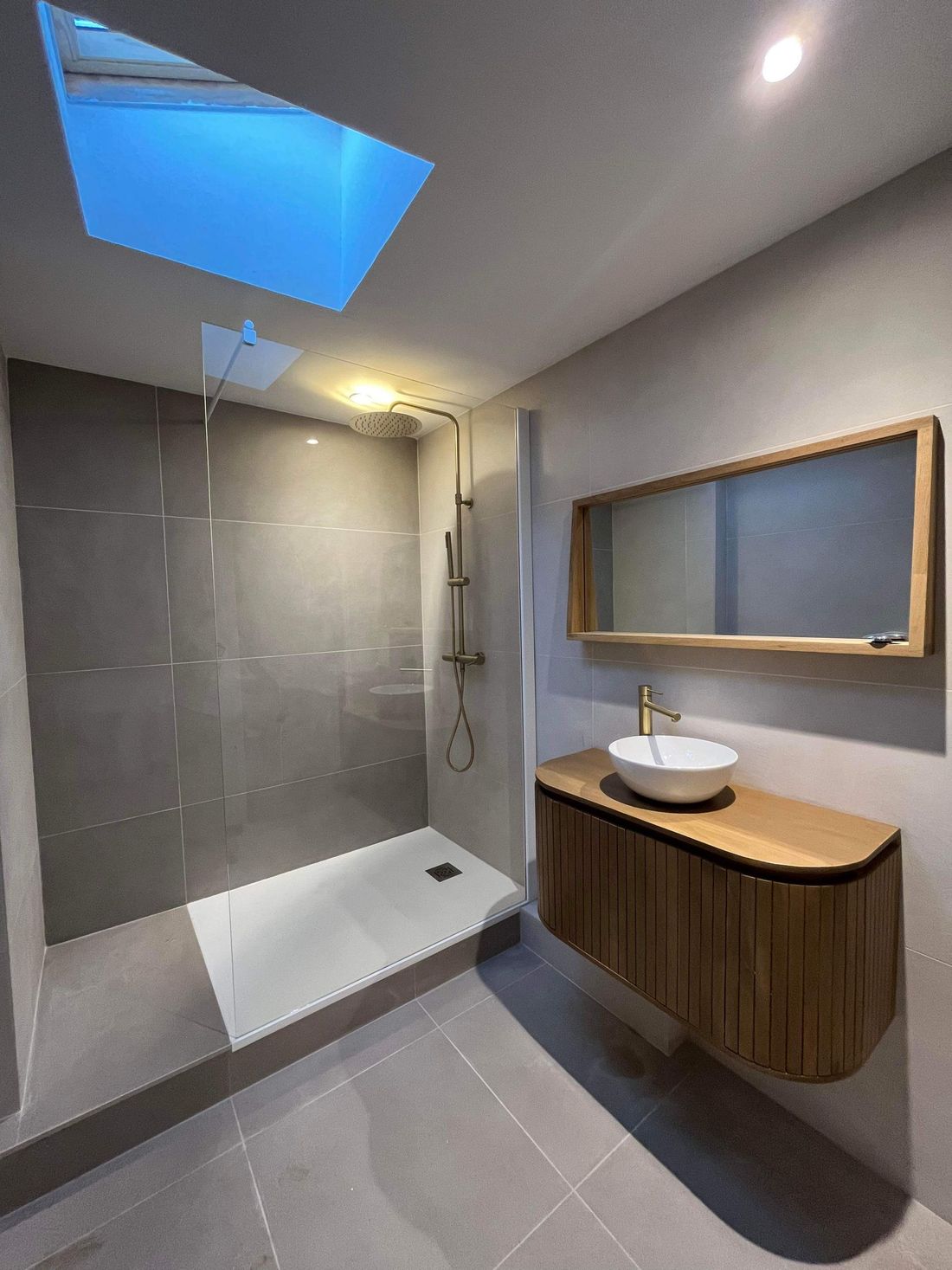 Salle de bains moderne avec douche à l'italienne, puits de lumière, carrelage gris et meuble lavabo en bois avec vasque ronde.