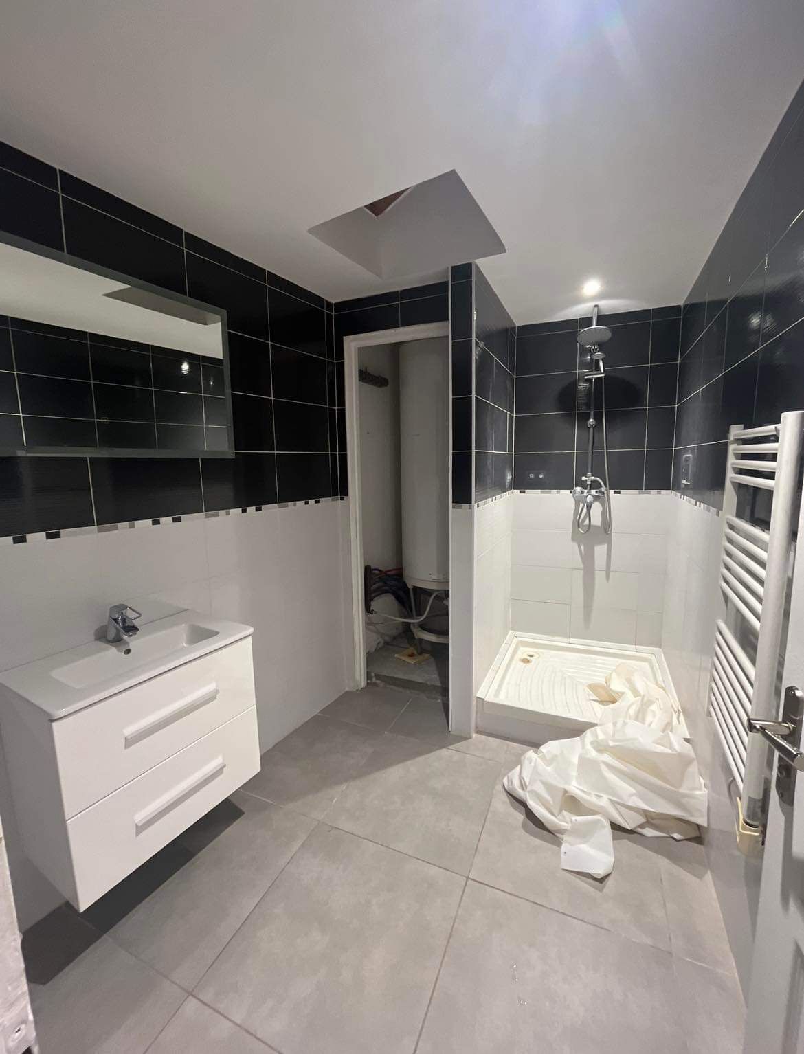 Salle de bains moderne avec murs carrelés en noir et blanc, lavabo, porte ouverte et linge au sol.