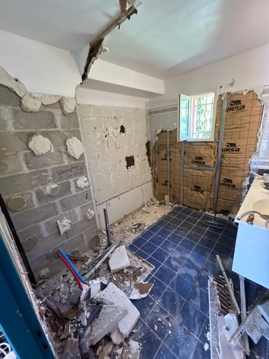 Pièce endommagée avec des pans de murs apparents, des débris au sol et une fenêtre cassée.