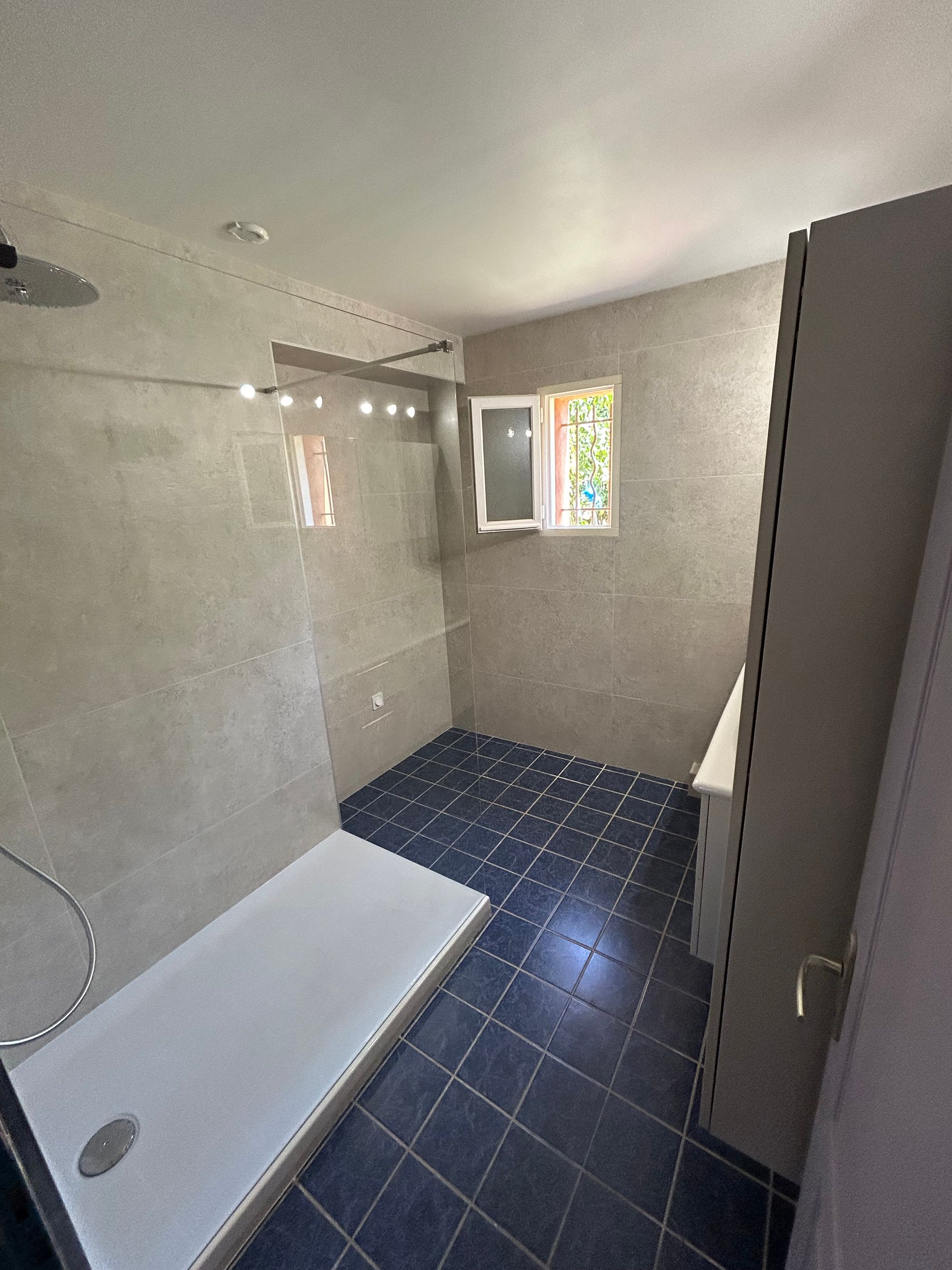 Petite salle de bains avec baignoire blanche, sol carrelé bleu foncé et une fenêtre laissant entrer la lumière.