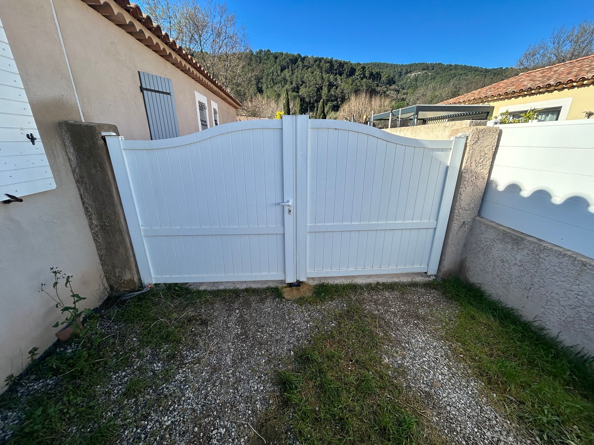 Un portail double blanc, entre des murs, ouvre sur une cour gravillonnée avec un centre gazonné.