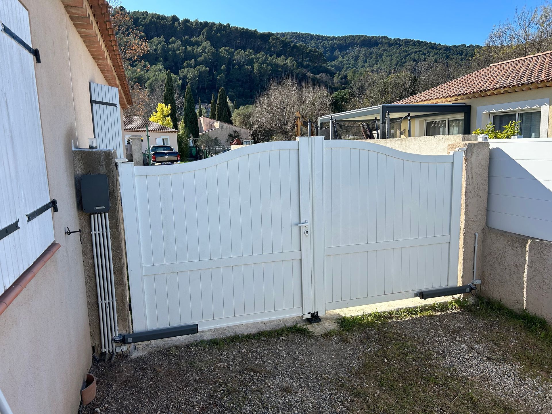 Portail double blanc donnant sur une allée, à côté d'une maison, avec des collines et des arbres en arrière-plan.