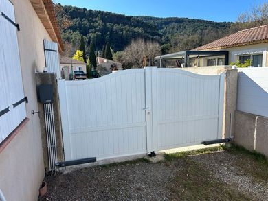 Portail double blanc donnant sur une allée, à côté d'une maison, avec des collines et des arbres en arrière-plan.