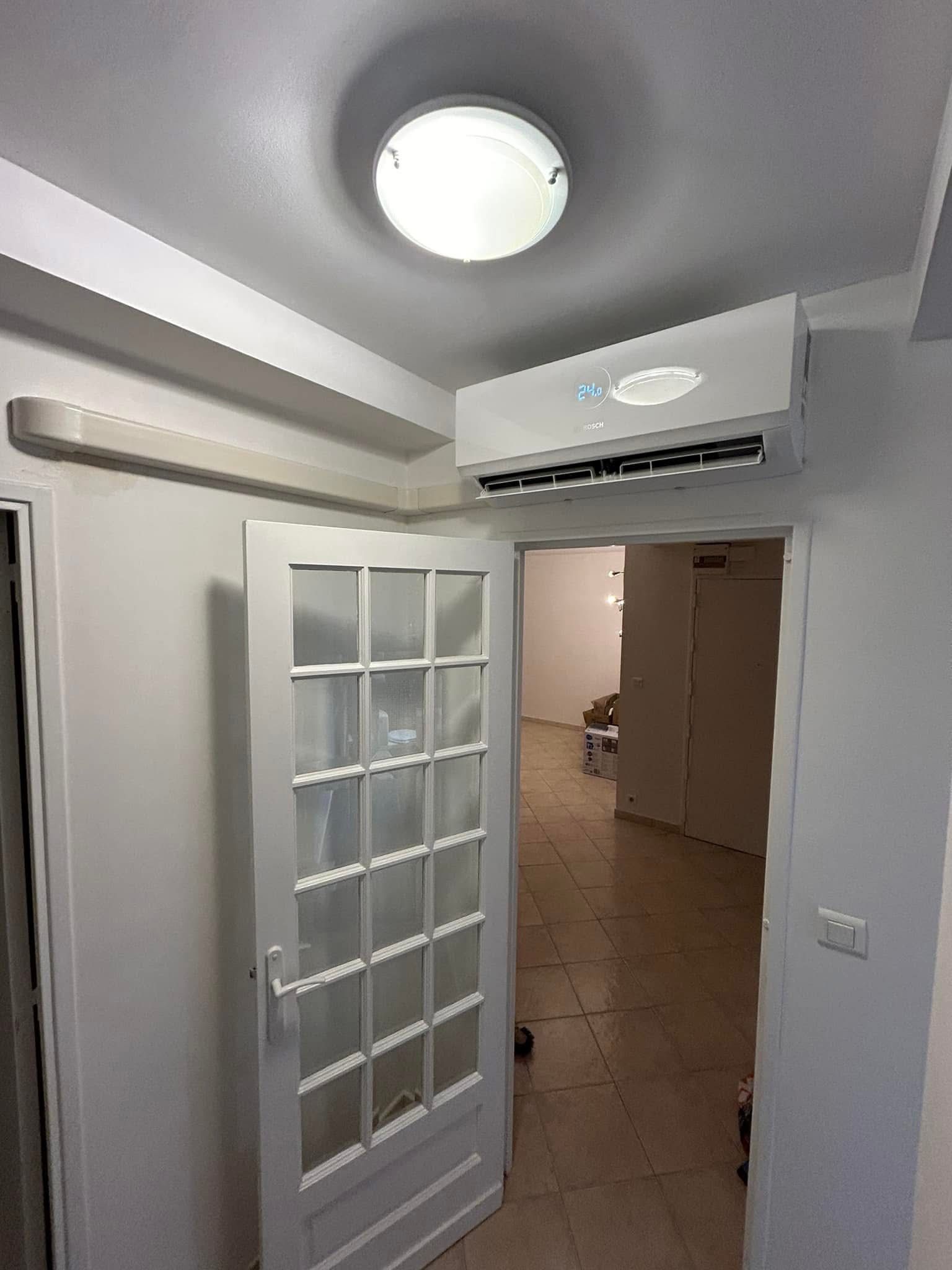 Porte vitrée ouverte dans un couloir blanc étroit avec un plafonnier et un climatiseur.
