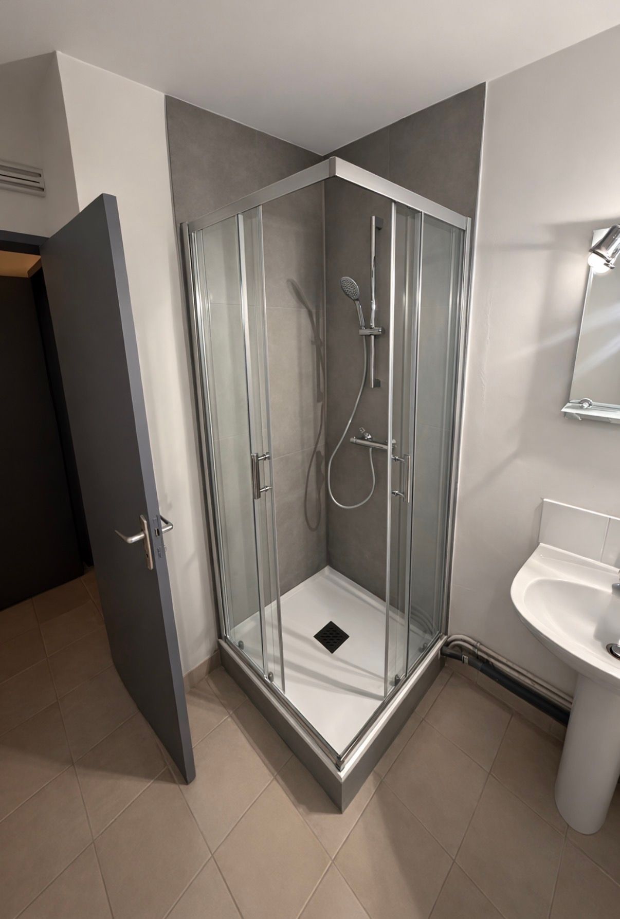 Cabine de douche moderne avec portes en verre, carrelage gris et lavabo blanc à côté.
