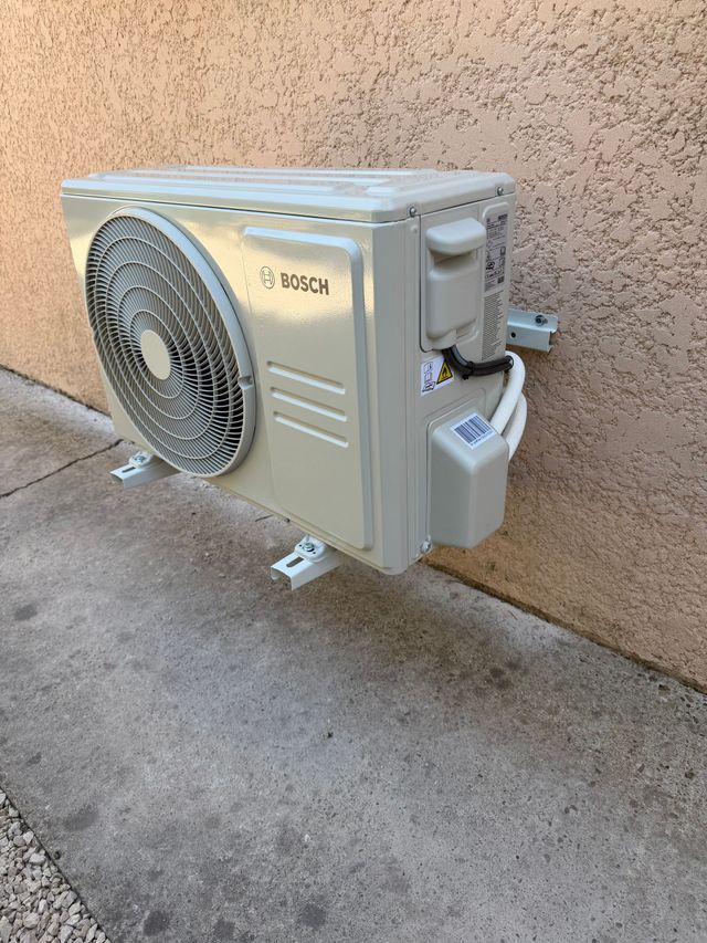 Unité de climatisation murale extérieure avec ventilateur, fixée sur un mur beige.