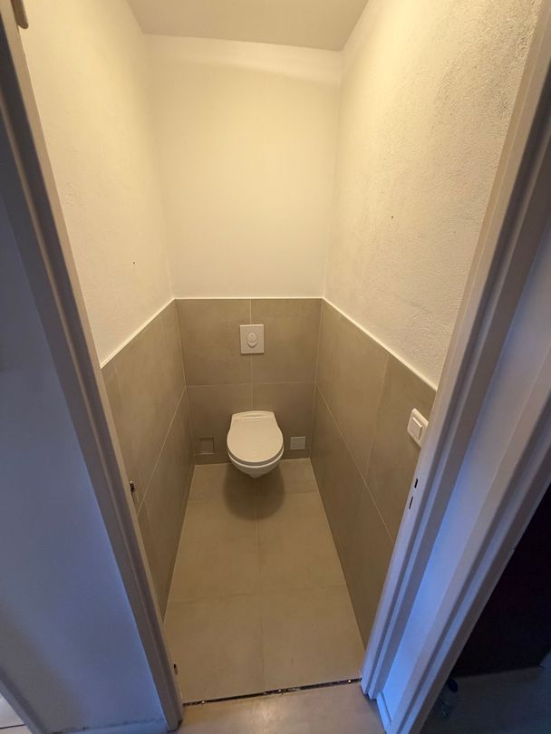 Petite cabine de toilettes étroite avec WC, murs beiges et vue depuis la porte ouverte.