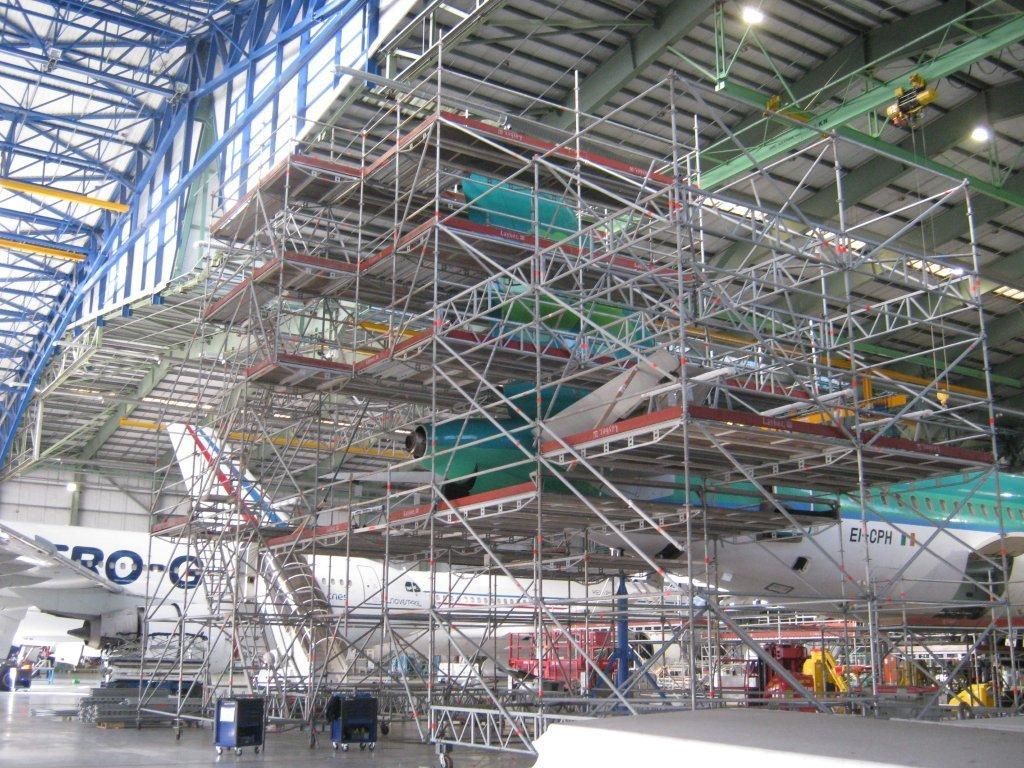 Un avion est entreposé dans un hangar sous un échafaudage. La structure métallique argentée, à plusieurs niveaux, est accessible par des escaliers. L'appareil est vert et blanc.