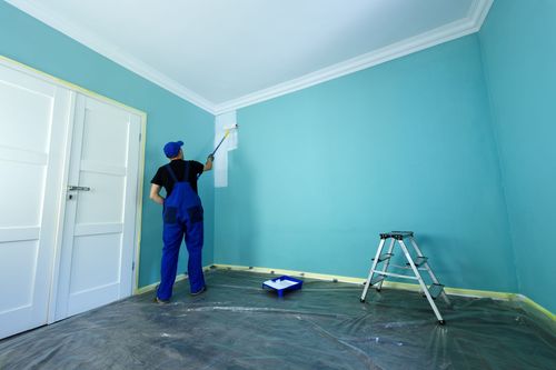 Un peintre en salopette bleue peint une pièce turquoise avec une échelle, des moulures blanches et une bâche au sol.