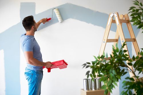 Peindre un mur blanc en bleu clair à l'aide d'un rouleau et d'un bac à peinture, à l'intérieur, avec une échelle à proximité.