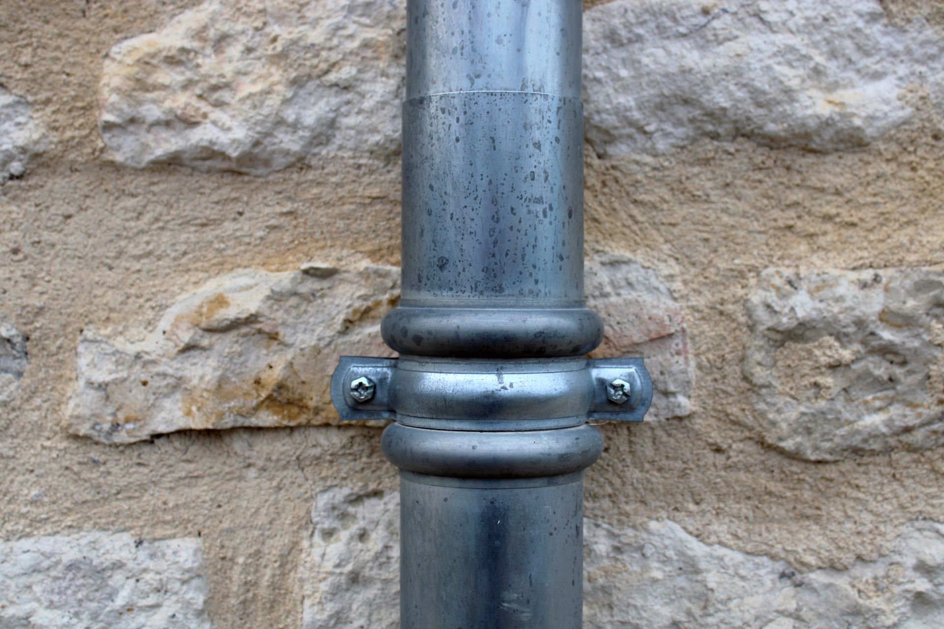 Collier de serrage métallique sur un tuyau gris vertical contre un mur en pierre beige.
