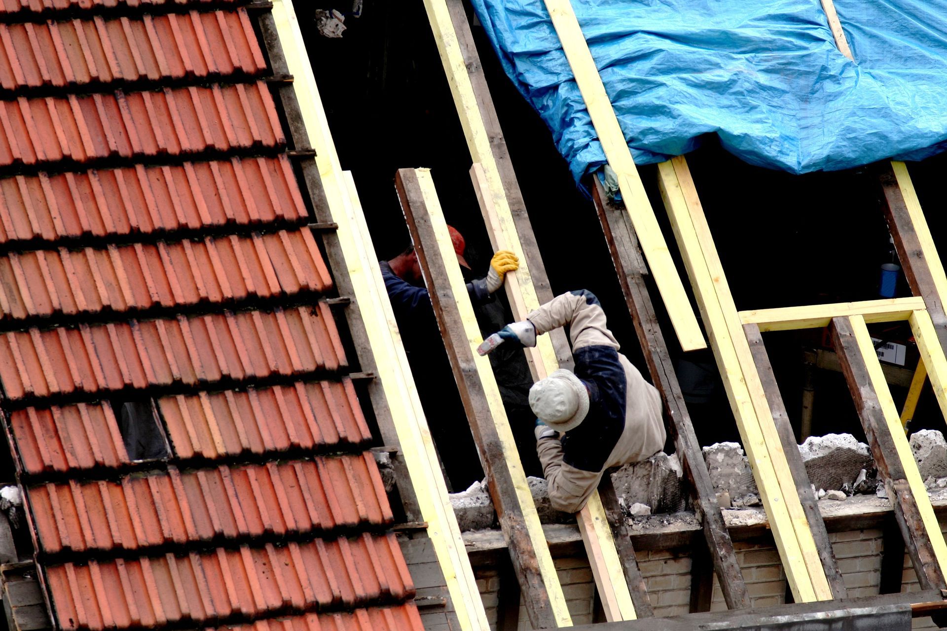 Un ouvrier travaille sur un toit en construction, à côté de tuiles rouges et d'une bâche bleue, parmi des poutres en bois.