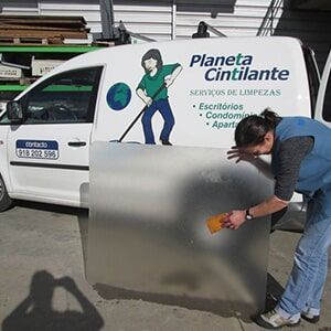 Mulher limpando um painel de vidro fosco ao ar livre, próximo a uma van de serviço de limpeza com o logotipo da empresa.