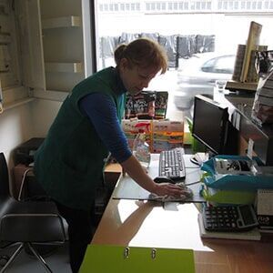 Mulher de colete verde limpando um balcão em uma loja; computador, teclado e outros itens visíveis.